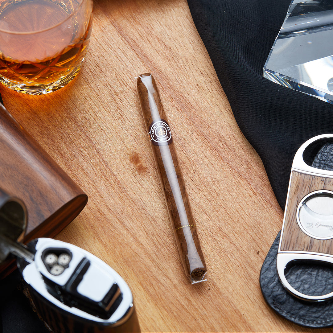 Montecristo Puritos Medium-Full Strength Cuban Cigarillo in its protective cellophane wrap, elegantly placed on a wooden surface alongside cigar accessories and a glass of whiskey.