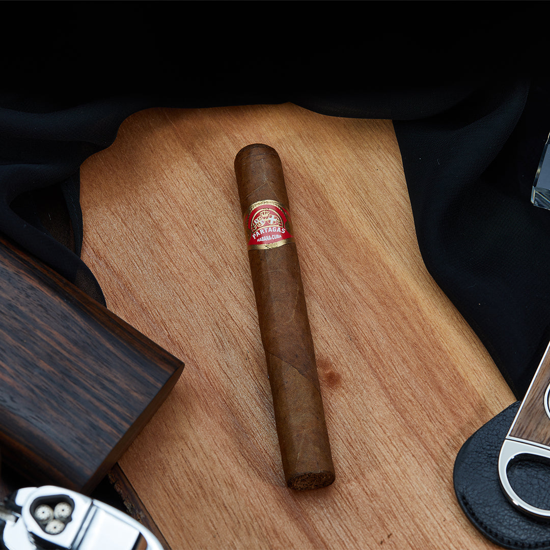 Partagás Mille Fleurs Cuban cigar with a red and gold band, placed on a wooden surface alongside a cigar cutter, lighter, and smoking accessories.