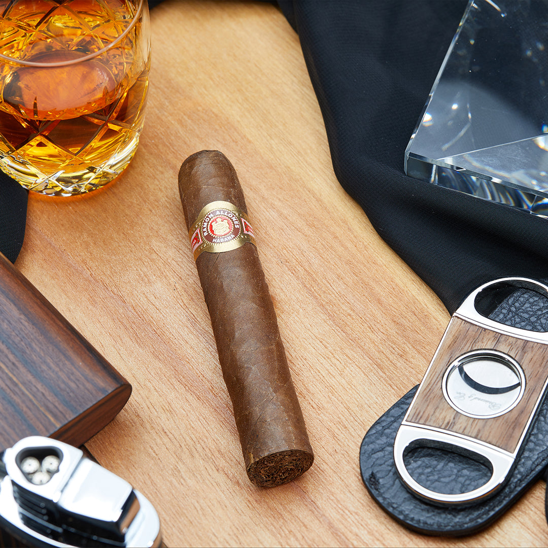 A Ramon Allones Specially Selected Cuban cigar resting on a wooden surface, accompanied by a whiskey glass, a cigar cutter, and other smoking accessories.