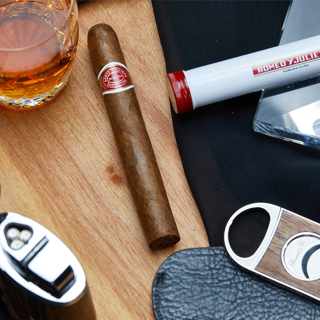 Romeo y Julieta No. 2 Cuban cigar resting on a wooden surface, alongside its aluminum tube, a whiskey glass, a cigar cutter, and a lighter, showcasing its iconic red and white band.