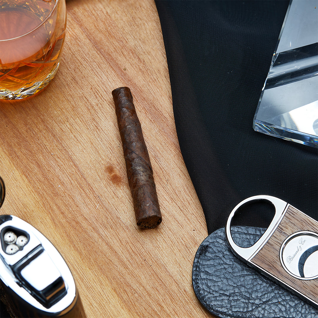 A Toscano Toscanello Medium-Full Strength Italian Cigarillo resting on a wooden surface, accompanied by a whiskey glass, a cigar cutter, and a triple-flame lighter, creating a refined and elegant smoking ambiance.