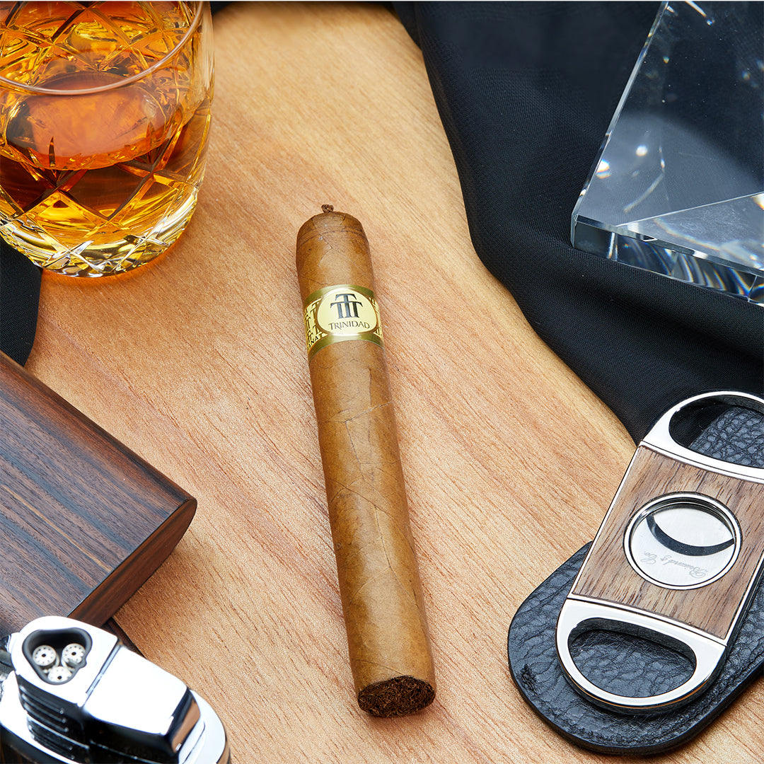 Trinidad Coloniales Cuban cigar resting on a wooden surface, accompanied by a whiskey glass, a cigar cutter, and a lighter, showcasing its golden band and signature pigtail cap.